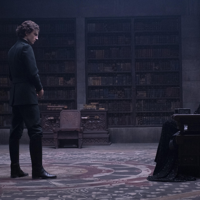 Un homme vêtu de noir se tient devant une femme elle aussi vêtue de noir assise dans une chaise massive en bois dans une salle dont les murs sont remplis de livres.