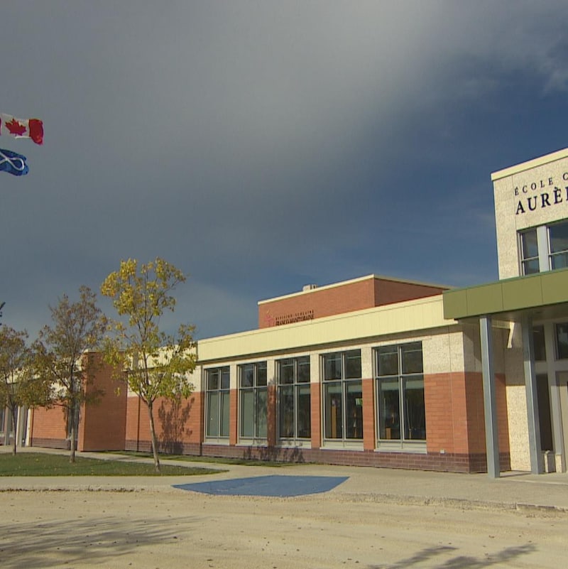 une école : on voit le bâtiment, un peu du stationnement et le mât, ainsi que les drapeaux canadiens et métis qui flottent. 