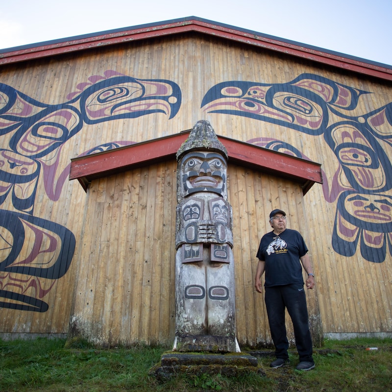 Charlie Mason pose à coté d'un totem traditonnel autochtone devant une maison en vois avec des peintures traditonnelles autochtones, à Klemtu, en Colombie-Britannique, en octobre 2023.