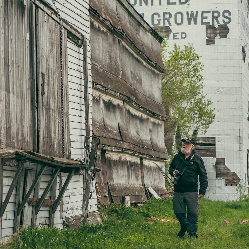 Gordon Goldsborough regarde un ancien silo à grain, avec une casquette et son appareil photo en bandoulière à Barnsley au Manitoba, début juin 2022.