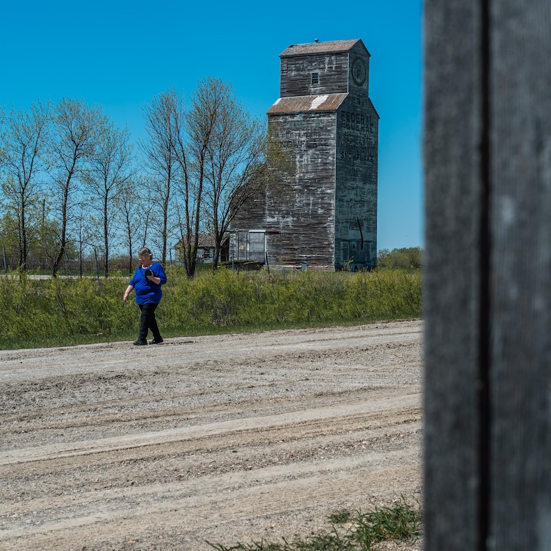 Jean McManus marche sur une route en gravier, elle tient son appareil photo dans une main, un silo à grain trône à l'arrière-plan, à Snowflake au Manitoba, début mai 2022.
