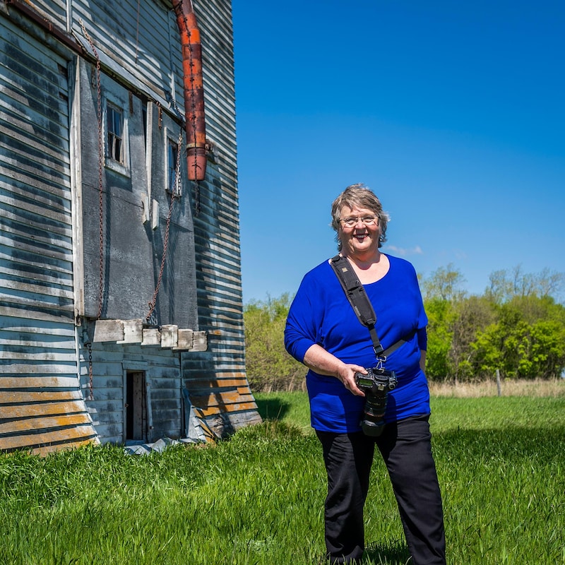 Jean McManus est devant un ancien silo à grain, dans les prairies, elle tient son appareil photo dans une main à Snowflake au Manitoba, début mai 2022.