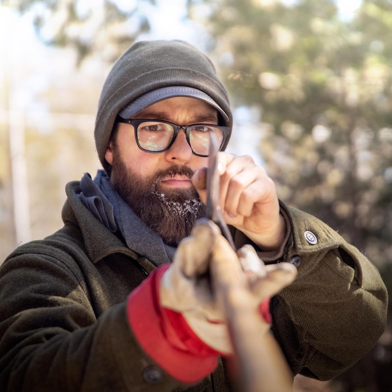 Un homme barbu à lunettes et tuque examine minutieusement la lame d'une hache dans les bois.