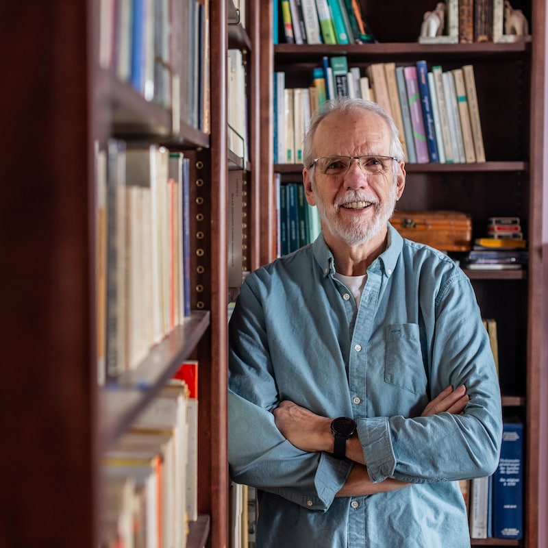 Un homme à la barbe blanche pose en souriant, les bras croisés, devant deux bibliothèques pleines de livres.