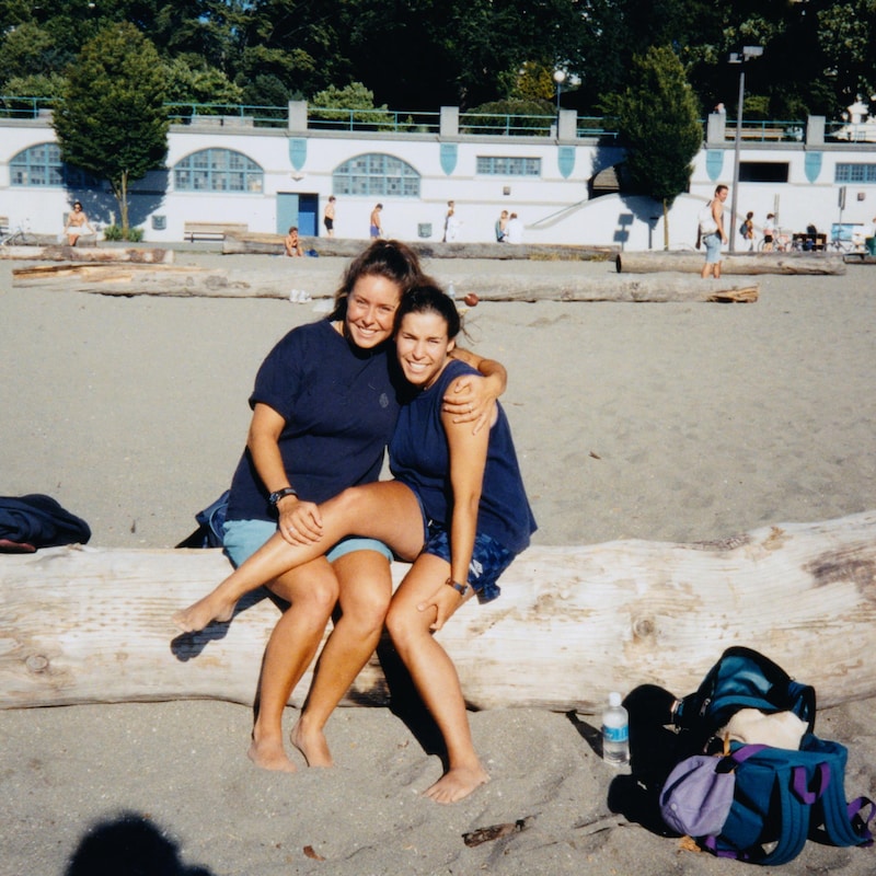 Joëlle et sa sœur se tiennent accolés sur un tronc d'arbre mort, sur une plage.
