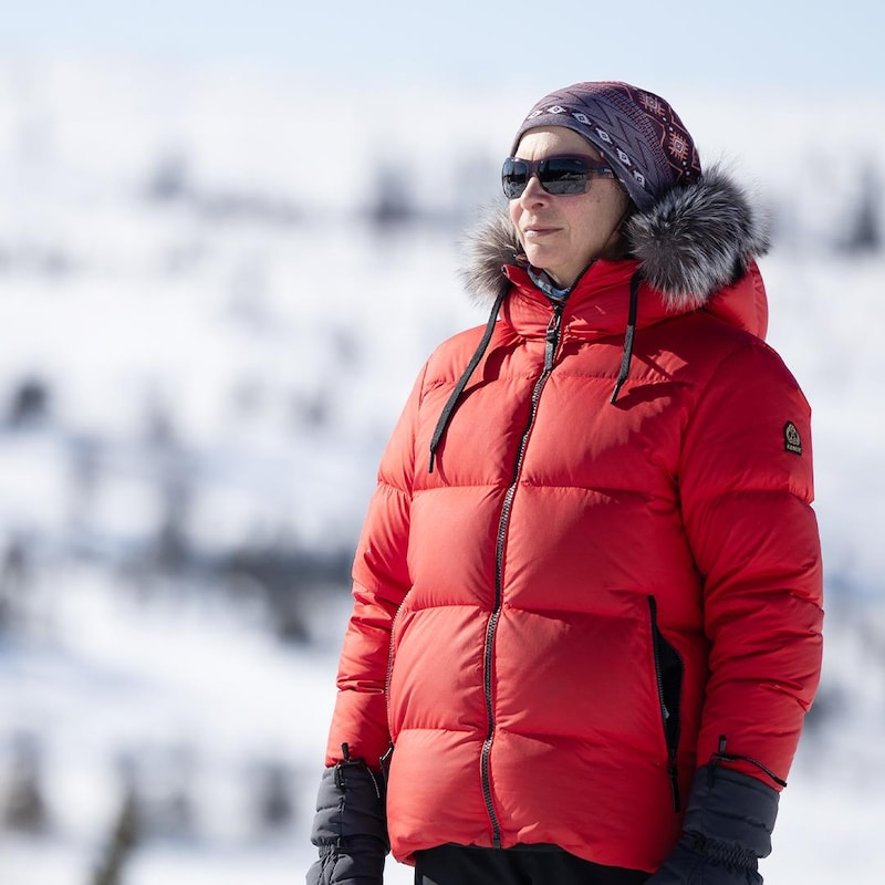 Une femme en manteau d'hiver rouge regarde au loin, devant une montagne enneigée.