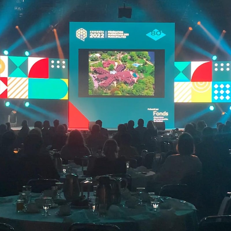 Une salle avec des gens assis à des tables qui regardent une présentation vidéo.