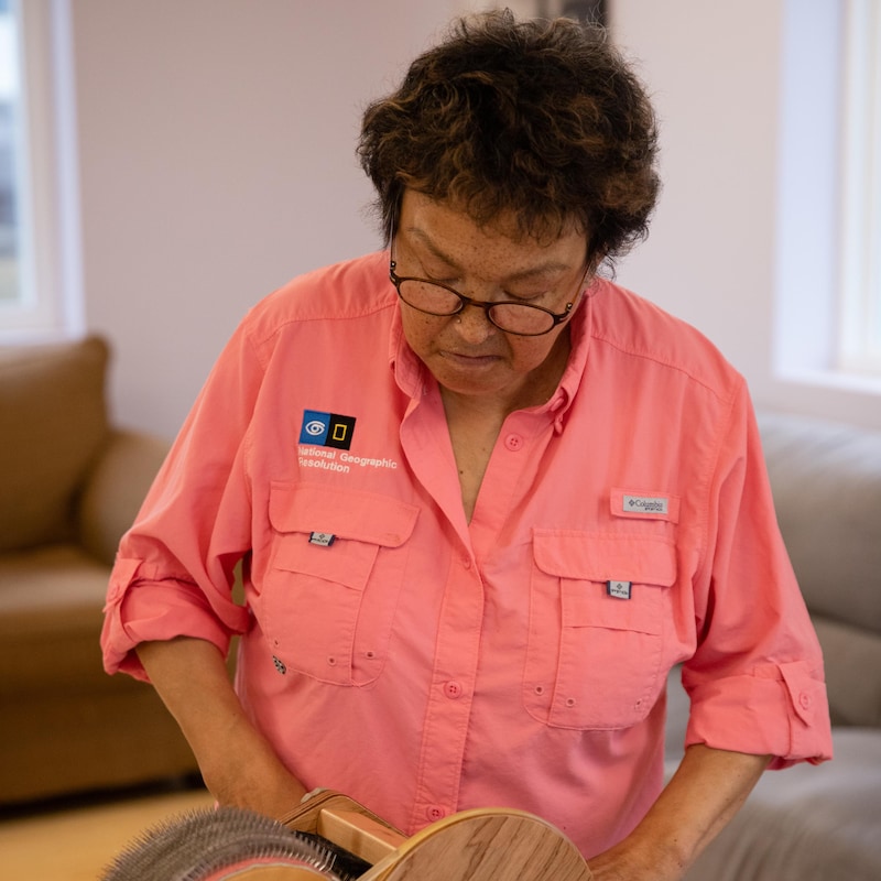 Annie est une femme dans la soixantaine. Elle porte une chemise de couleur corail et manipule une cardeuse.