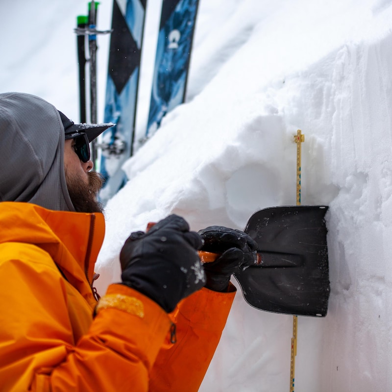 Un homme en habit d'hiver utilise une courte pelle pour gratter la neige afin de mesurer celle-ci. En arrière-plan, ses skis piqués dans la neige.