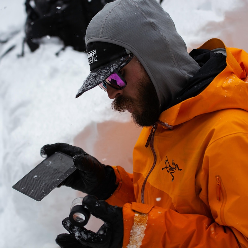 Un homme en habit de neige tient dans ses mains gantées une loupe et une plaquette pour observer la grosseur des flocons de neige.
