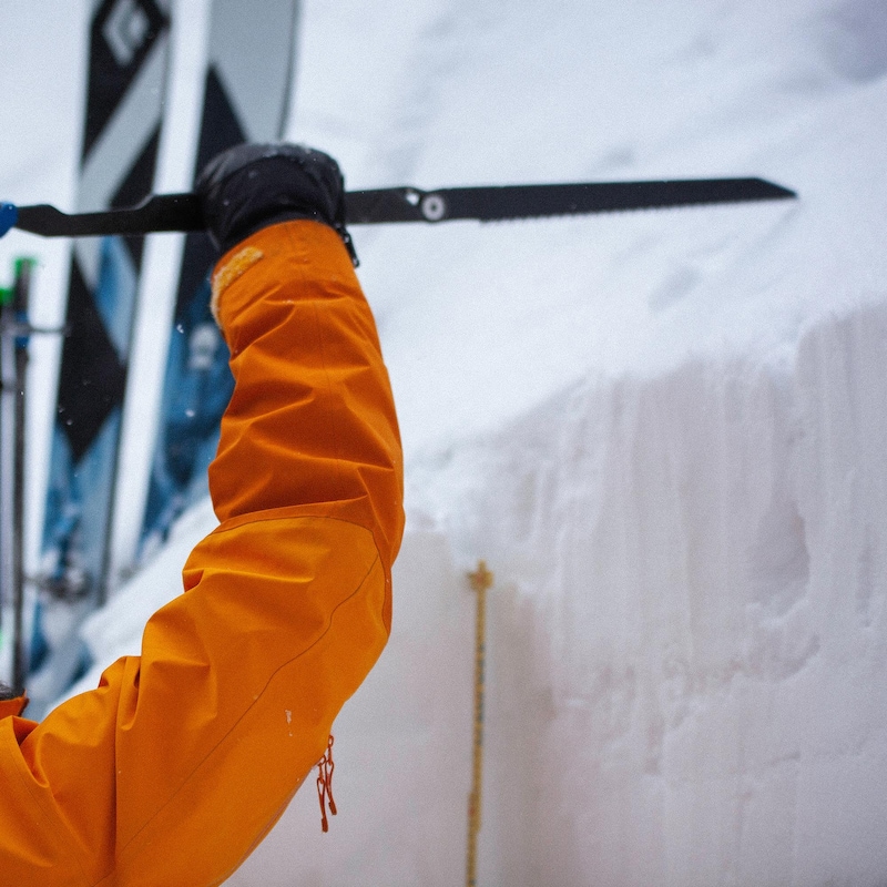 Un homme les bras en l'air tient un bâton pour mesurer la quantité de neige. En arrière-plan, ses skis plantés dans la neige.