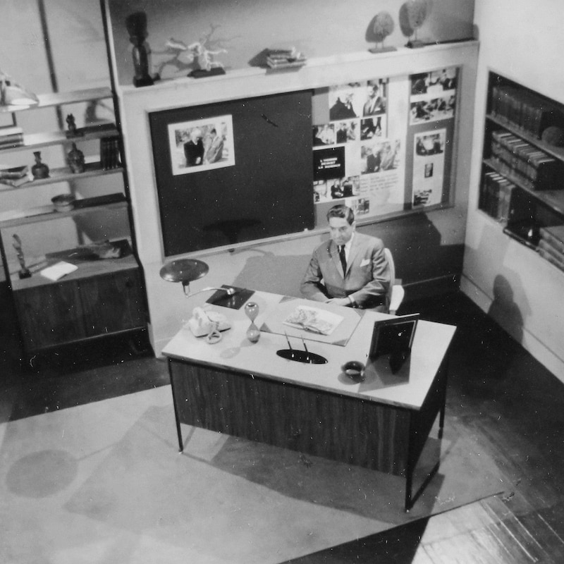 Fernand Seguin derrière un bureau avec un tableau derrière lui dans le décor de l'émission.
