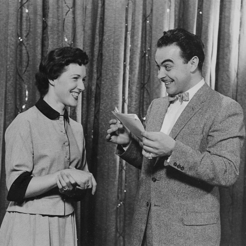 Sur une scène, Henri Bergeron, feuilles à la main, regarde une concurrente en souriant.