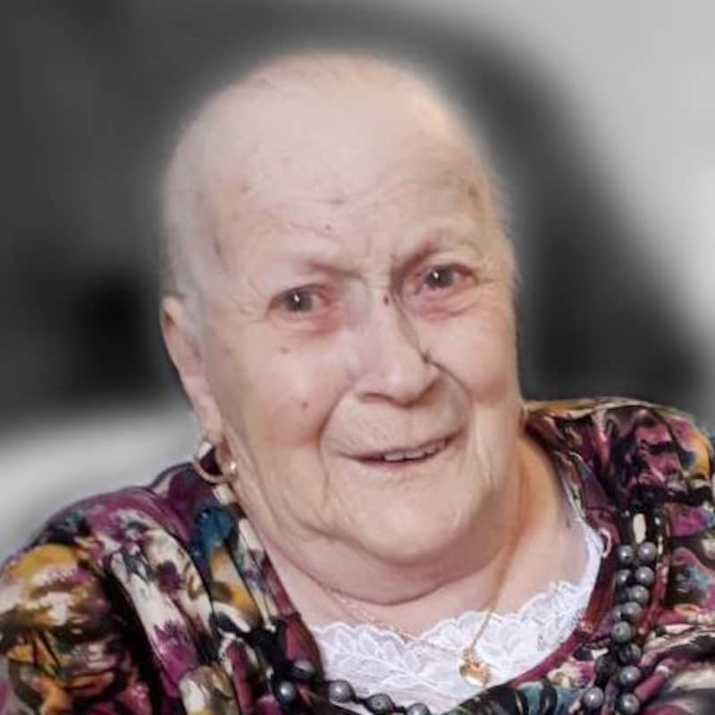 Madame Brassard est assise dans un fauteuil et réagit à ce que le ou la photographe lui dit. Elle porte une blouse à motifs mauves.