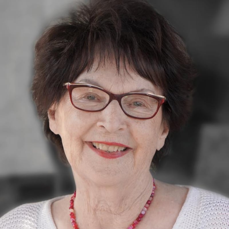 Madame Baaudin sourit pour la photo. Elle porte du rouge à lèvres et un collier de teinte rouge. Elle a les cheveux noirs et des lunettes brunes.