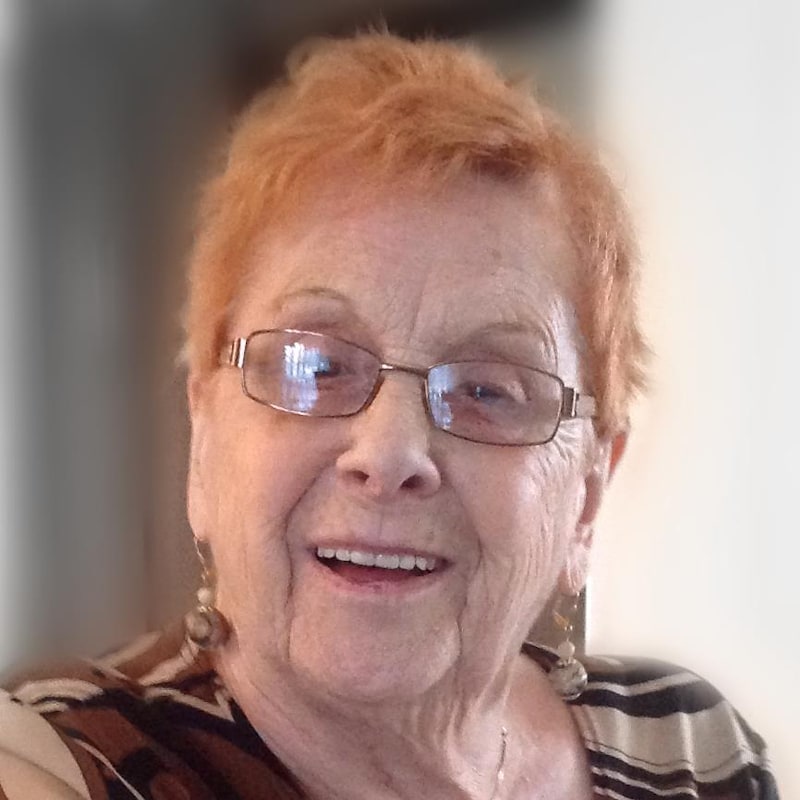 Madame Blais sourie à la caméra. Elle a les cheveux teints en roux, porte des lunettes et des boucles d'oreilles beiges.