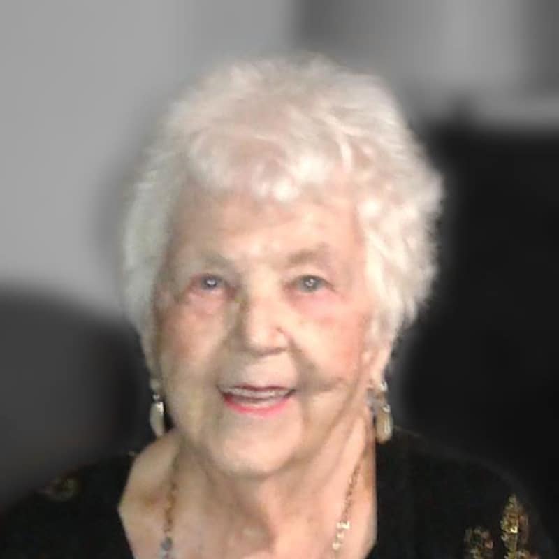 Simone regarde l'objectif d'un visage amusé. Elle porte un collier long et est vêtue de noir.