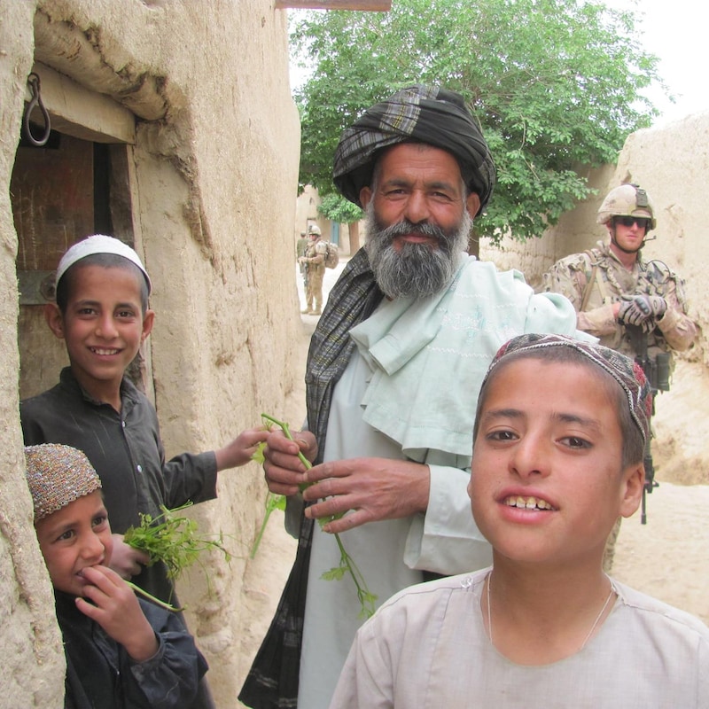 Trois enfants Afghans souriants et un adulte regardent la caméra. Un soldat canadien se tient derrière eux.