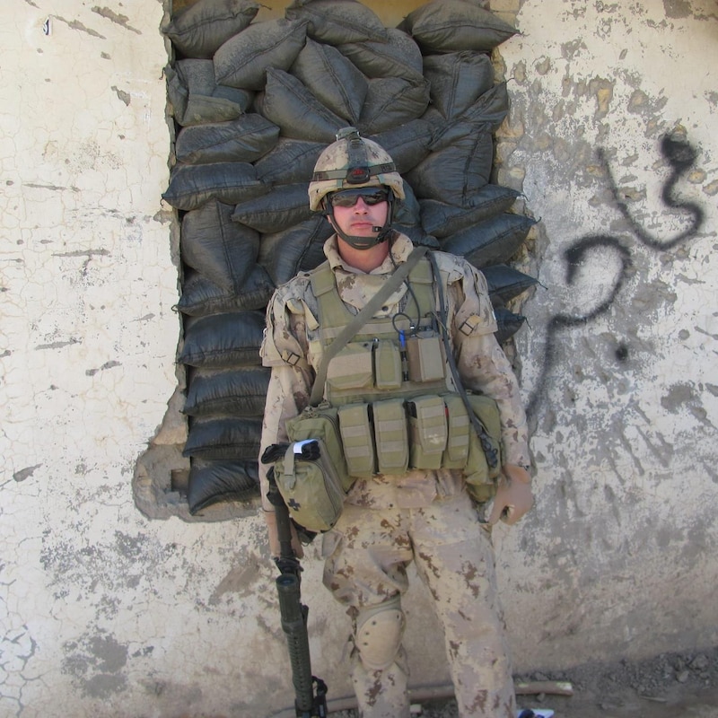 Patrick Lemay armé et en habit militaire se tient devant un mur où les fenêtre ont été remplies de sacs de sable.