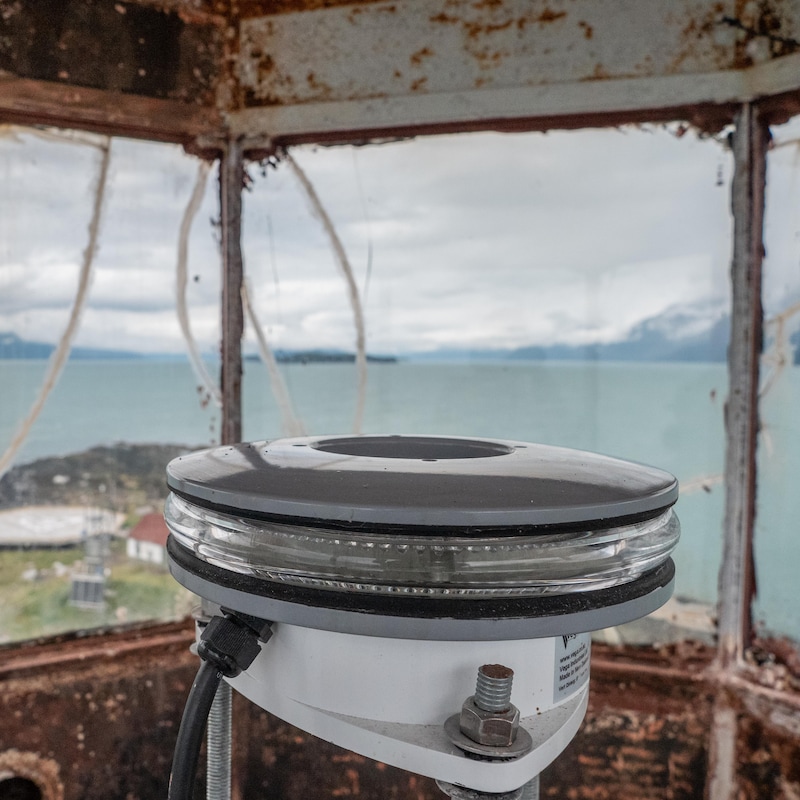 Système lumineux du phare avec dans le fond la vue depuis le phare qui donne sur le rocher et la côte au loin, en Alaska, juillet 2022.
