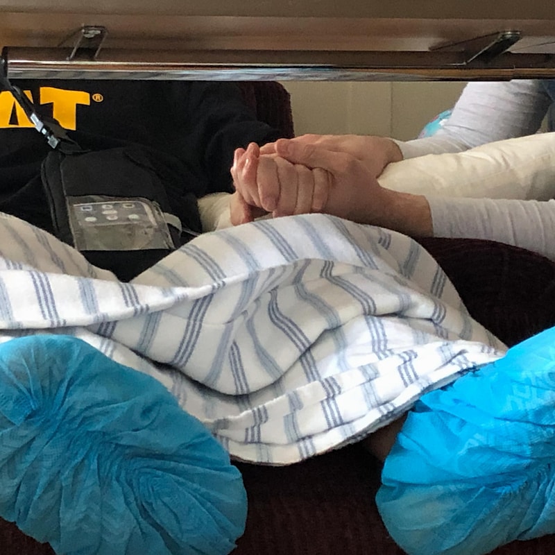 Un homme malade étendu sur une chaise tient la main d’un autre homme.