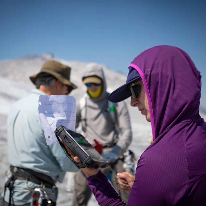 Une femme regarde un appareil pendant que deux hommes sont debout en arrière-plan, sur un glacier.