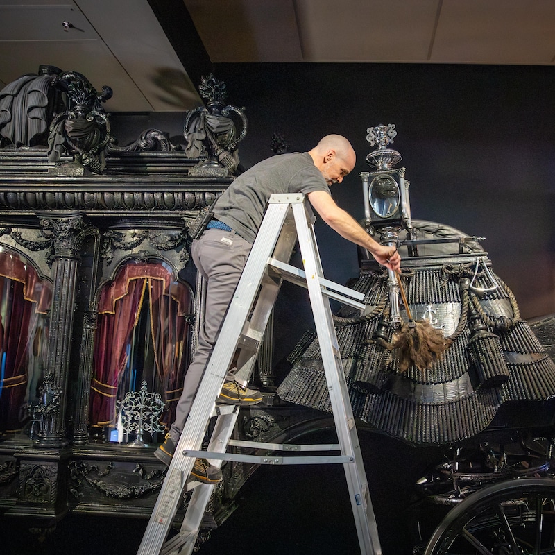 Un employé du Musée de la civilisation, debout sur un escabeau, utilise un plumeau pour nettoyer les éléments décoratifs à l'arrière d'un imposant corbillard hippomobile noir.