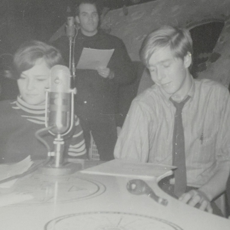 Claude Quenneville assis devant un micro à une table, consultant ses feuilles. Près de lui, Odette Brassard et Jean Fortier.