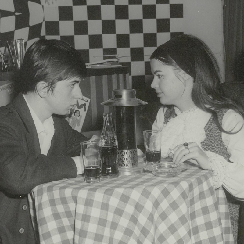 Deux étudiants incarnés par Louise Portal et Ghislain Tremblay assis à une petite table ronde dans un restaurant.