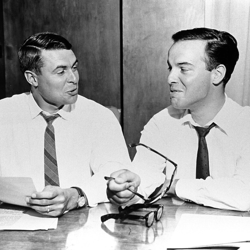 Dans un studio de radio, les animateurs Richard Garneau et Pierre Nadeau rigolent en se regardant, assis derrière un micro suspendu.