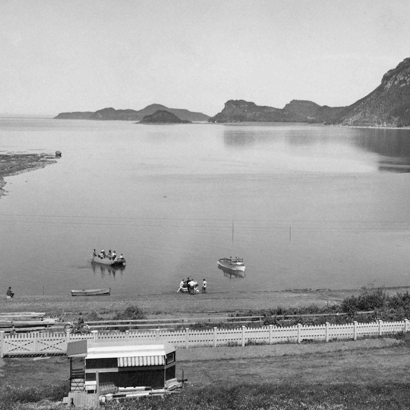 Photo d'archives en noir et blanc : des chaloupes sur le fleuve avec des montagnes au loin.