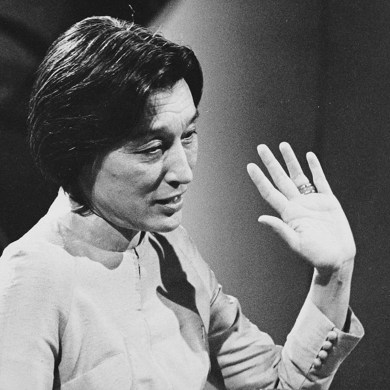 Dans un studio de télévision, l'écrivaine Han Suyin, levant le bras lors d'une entrevue. Un micro de table est posé devant elle.