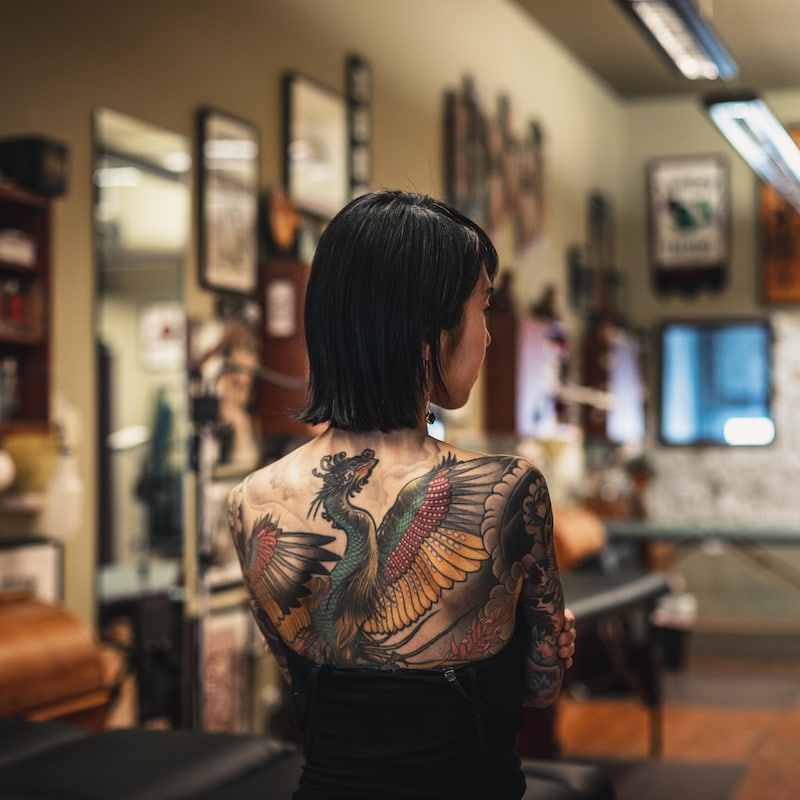Une femme de dos dans un salon de tatouage exhibe un dragon coloré dessiné sur son dos.