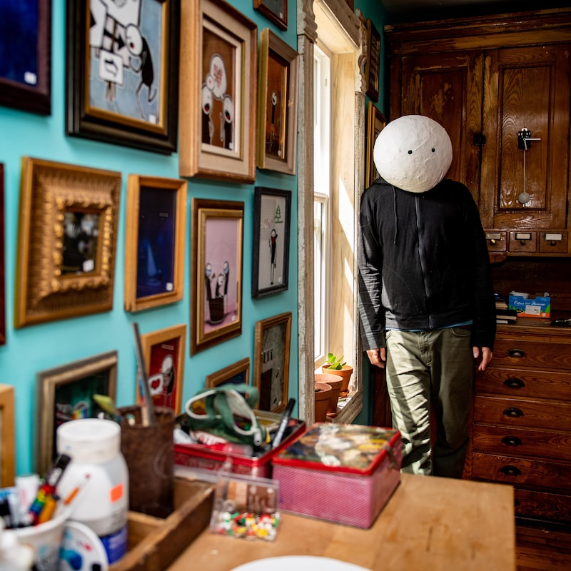 Ultra Nan avec un masque sur la tête dans son atelier. Au mur, on retrouve des dizaines de petits cadres avec ses oeuvres. 