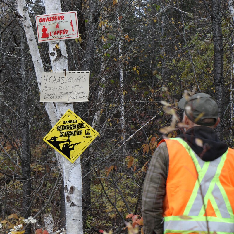 Sur un tronc d'arbre, plusieurs panneaux indiquant la présence de chasseurs. Un homme de dos regarde.