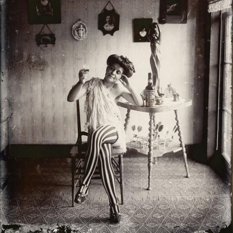 Une femme légèrement vêtue, assise sur une chaise et accoudée contre une table, lève un petit verre.