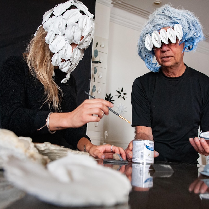 Une femme et un homme portant des masques formés de vulves trempent un pinceau dans un pot de peinture blanche.
