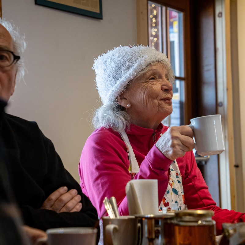 Deux aînés à la table d'un restaurant. À gauche un homme parle avec son voisin (qui n'est pas sur la photo). À droite, une femme sourit, une tasse de café à la main. 