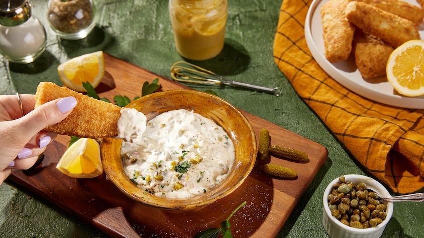 Un bol de sauce tartare servi avec des quartiers de citron, des câpres, des cornichons et des bâtonnets de poisson.