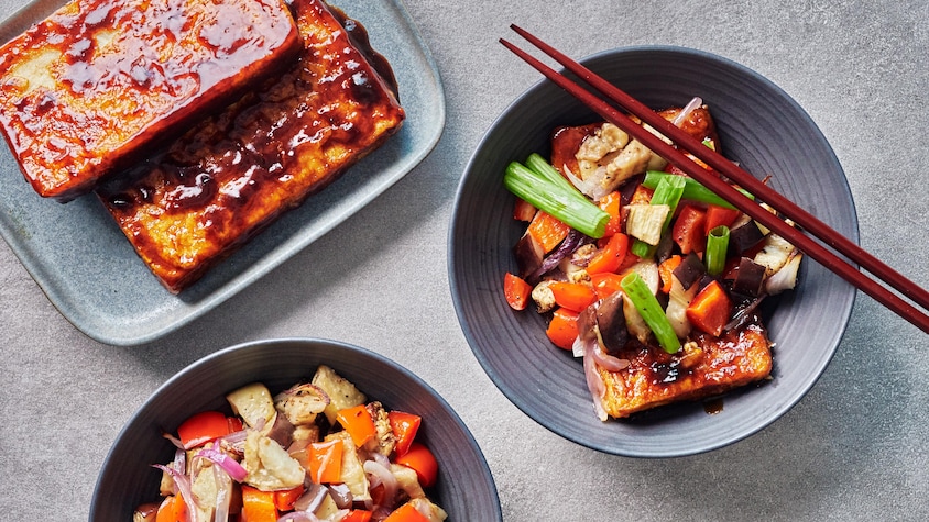 Des pavés de tofu caramélisés dans un plat de service et deux assiettes contenant des morceaux de tofu et des légumes.