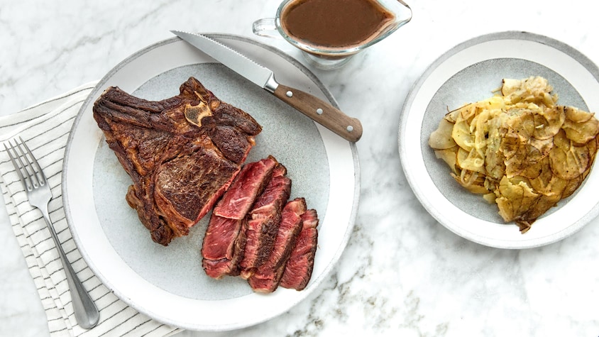 Sur un comptoir, il y a une assiette contenant des pommes de terre boulangères, une autre assiette contenant du bifteck de côte de bœuf ainsi qu'un saucier rempli de sauce brune.
