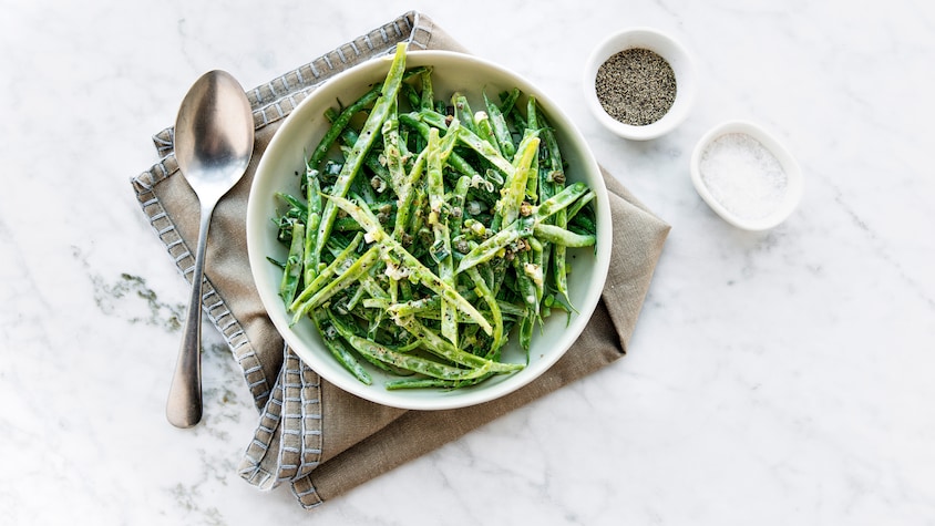 Un bol rempli d'haricots verts avec à ses côtés une cuillère, un bol de poivre et un bol de sel.