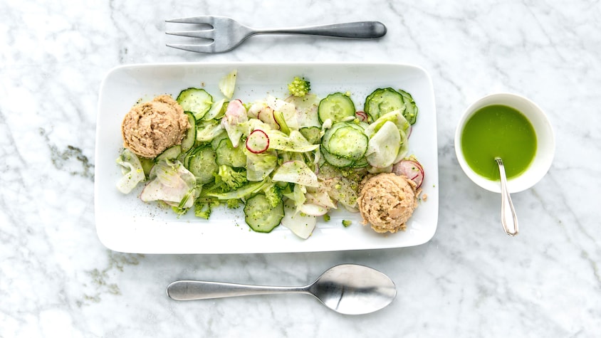 Une salade de légumes dans une assiette de service avec deux boules de thon avec à ses côtés un bol de vinaigrette verte.