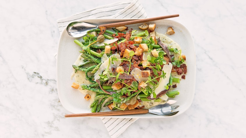 Des asperges avec tous les ingrédients du salade César dans une assiette de service.