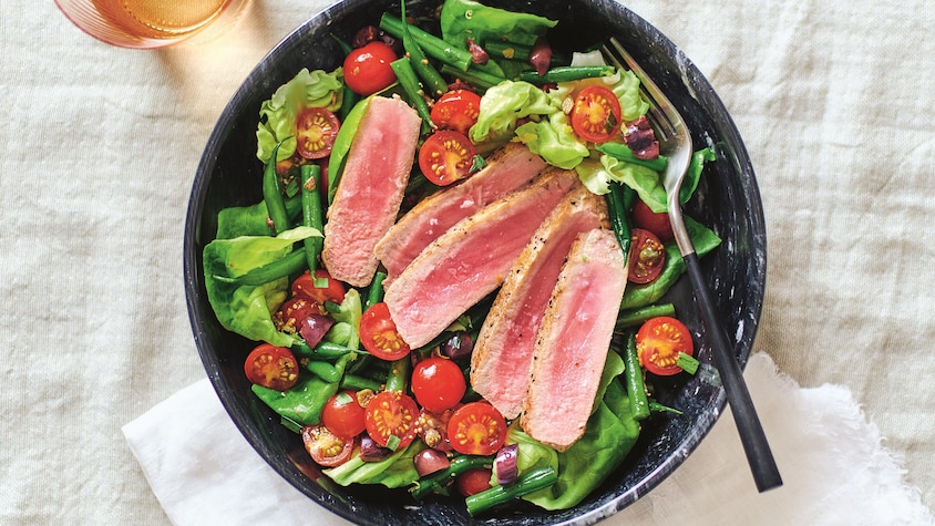 Des tranches de thon frais sur une salade verte composée de haricots verts, de tomates cerises, ainsi que des feuilles de laitue Boston.