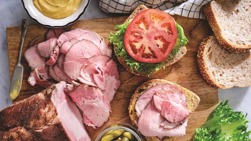 Sur une planche à découper, il y a du rôti de porc tranché et une tranche de pain garnie de viande. Il y a aussi une autre tranche de pain garni de laitue et d'une tranche de tomate pour faire un sandwich.