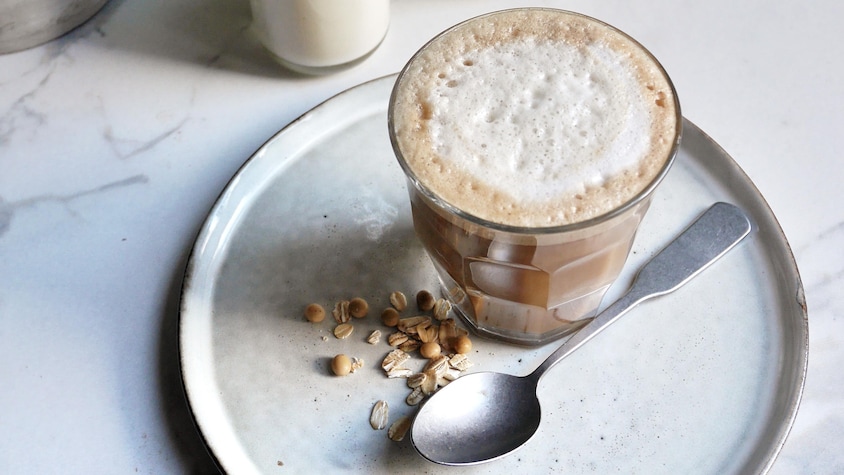 Un verre de boisson soya et avoine bien moussée pour le café, dans une assiette, à côté de grains de soya et d'avoine, et d'une cuillère à café.