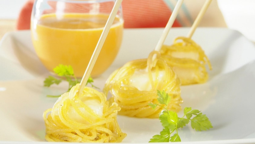 Trois fondues parmesan enveloppés de spaghettis disposés dans une assiette blanche aux côtés d'un verre à cocktail de coulis jaune.