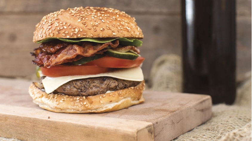 Un burger garni de fromage, de tomates, de laitue et de bacon posé sur une planche en bois.