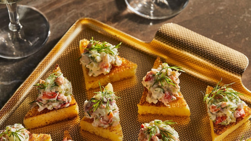 Plusieurs triangles de pain grillé recouverts d’une préparation au homard et d’une feuille d’aneth fraîche dans un plateau doré avec deux coupes de vin blanc.